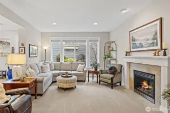 The living room feels bright and inviting with large windows that draw in natural light and frame the gardens outside. The gas fireplace is elevated by a beautifully detailed mantel and a recessed art light above, creating a refined focal point that anchors the entire space.