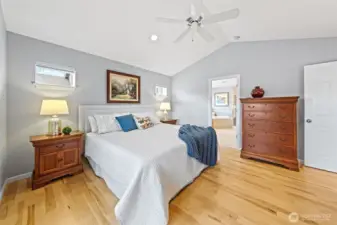 The Primary Suite feels bright and serene with vaulted ceilings and two elevated accent windows that bring in soft natural light while preserving privacy. A ceiling fan provides added comfort and cooling, while the open flow into the en-suite bath enhances the room’s airy, restful retreat feel.