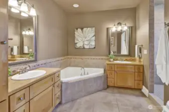 Attached primary bath features a corner jetted tub, double sink vanity, & a tiled walk-in-shower with two shower heads.