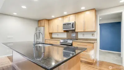 Modern kitchen with natural toned cabinetry. Spacious island features a sink, dishwasher, outlet, and plenty of storage—perfect for meal prep and casual gatherings.