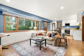 Bright and inviting open-concept living space with a full kitchen, dining, and cozy seating area, all framed by expansive windows that bring in natural light and capture views of the surrounding greenery.