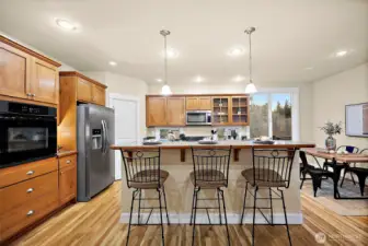 Island with bar seating, in-wall oven, side-by-side refrigerator, wood cabinetry and hardwood floors