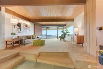 This spacious living room captures the essence of Northwest modern design, framed by sweeping views of Puget Sound and the Olympic Mountains. Warm wood ceilings with exposed beams, expansive floor-to-ceiling windows, and a seamless indoor-outdoor connection create a serene backdrop for everyday living and effortless entertaining. Natural light pours across the cozy seating area, highlighting the room’s timeless architectural character and inviting atmosphere. A rare blend of iconic design and breathtaking vista