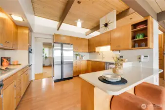 Bright modern kitchen featuring warm wood cabinetry, exposed beam ceilings, and newly added skylights that flood the space with natural light. Spacious layout with stainless steel double-door refrigerator, light countertops, and an L-shaped design with peninsula seating. Open shelving and stylish finishes add charm, while the kitchen flows seamlessly into adjacent living areas—perfect for everyday living and entertaining.
