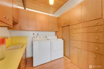 Well-appointed laundry room offers exceptional storage and everyday convenience with its extensive cabinetry, generous counter space, and built-in utility sink. Warm wood finishes and a paneled ceiling create a clean, cohesive look, while the full-size washer and dryer sit neatly within a highly functional layout. Thoughtful organization and timeless design make this a practical and inviting workspace for household tasks.