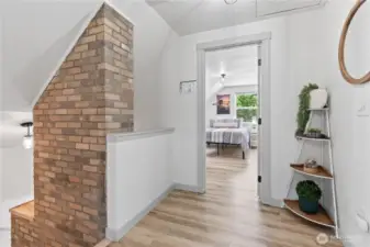 This inviting upstairs hallway features a charming brick accent chimney and leads into a bright, spacious bedroom. The open walkway is adorned with thoughtful decorative shelving and warm-toned flooring, creating a natural transition between the home’s upper-level living spaces.
