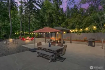 Dreamy outdoor oasis surrounded by towering evergreens! This twilight photo captures a beautifully lit backyard featuring a cozy fire pit, outdoor string lights, and multiple seating areas perfect for hosting guests or enjoying quiet evenings. The spacious patio includes a large dining table with an umbrella, ideal for al fresco dinners or summer BBQs. The peaceful forest backdrop and ambient lighting create an unforgettable setting.