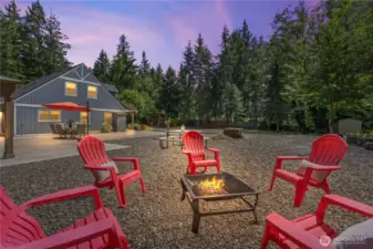 Twilight magic meets modern comfort in this beautifully staged fire pit area with red Adirondack chairs. The soft glow of the fire creates a cozy, welcoming atmosphere with the farmhouse-style home's lights adding warmth in the background. This gravel entertaining area is perfect for late-night conversations, roasting s’mores, or simply relaxing under the stars.