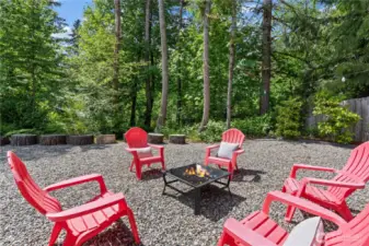 Bright and inviting, this forest-framed fire pit space is ready for unforgettable outdoor moments. Surrounded by mature trees and greenery, the vibrant red Adirondack chairs encircle a square fire feature, offering a perfect place to gather with friends and family. Log stump seats and string lights add rustic charm and functionality to this thoughtfully designed outdoor zone.