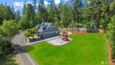 A stunning aerial view reveals the expansive backyard, complete with a fully-fenced grassy lawn, professional landscaping, and a dedicated play area with swings and a slide. The stylish paver pathway leads to the charming modern farmhouse, framed by mature trees and natural privacy. Perfect for families who value outdoor living, safety, and space to roam.