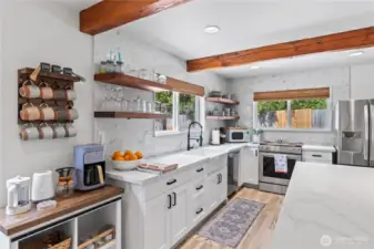 Enjoy modern convenience and rustic charm in this beautifully designed kitchen. Featuring a custom coffee station, a farmhouse-style apron sink, and stunning wood open shelving, this space blends functionality with personality. Natural light pours in through dual windows, accentuating the bright white cabinetry, textured backsplash, and warm wooden ceiling beams.