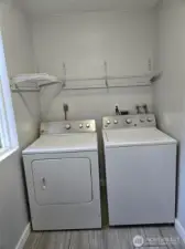 Convenient main-level laundry space features a side-by-side washer and dryer set, updated tile flooring, and upper wire shelving for added storage. A window adds natural light to brighten laundry days.