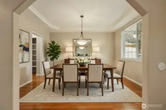 The formal dining room is framed by a graceful tray ceiling with crown detailing, creating a sense of height and architectural sophistication. Rich hardwood floors flow seamlessly throughout, while natural light from the oversized window enhances the warmth and openness of the space — ideal for both intimate dinners and larger gatherings.