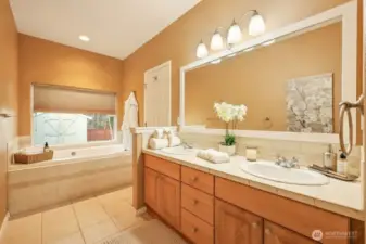The spacious primary bath offers a spa-inspired retreat with a double vanity, abundant cabinetry, and generous tile flooring. A deep soaking tub sits beneath a sunlit window, creating the perfect place to unwind. Warm finishes and thoughtful layout combine comfort, functionality, and timeless appeal.