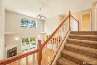 A stunning open-to-below design creates breathtaking volume and architectural presence. Sunlight pours through upper and lower windows, illuminating the hardwood floors and cozy fireplace below. The staircase, accented with rich wood railing and classic white spindles, adds refined craftsmanship and visual drama, making this home feel both grand and inviting.