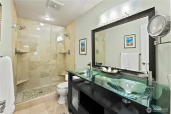 beautifully upgraded primary bathroom offers a spa-like experience with high-end finishes and thoughtful design. The spacious walk-in shower features floor-to-ceiling tile, a built-in bench, and frameless glass doors for a modern, open feel. The double vanity with vessel sinks, glass countertops, and rich espresso cabinetry provides both style and storage, complemented by sleek contemporary lighting.