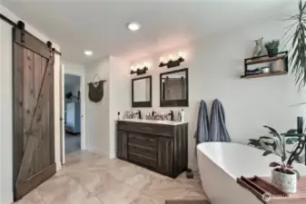 This photo showcases a master bathroom that incorporates a modern and stylish design element – a barn door leading to the bathing area, specifically featuring a free standing soaker tub. This combination of rustic barn door aesthetics with the contemporary look of a glass tub creates a unique and appealing space.