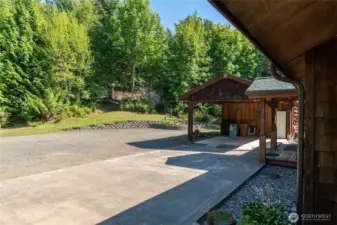 View from ramp of covered porch. Covered front entry and carport on right. Woodshed behind carport. Upper garage/workshop visible through the trees. Access upper garage with a circle driveway or center pathway.