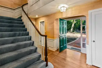 The gracious front entry welcomes you with timeless charm, setting the tone for the home’s warm and classic interiors. Natural light spills through the open front door, casting a glow on hardwood floors and illuminating the curved stair rail—a quiet invitation to explore the upper stories. Traditional paneling and millwork reflect the home’s heritage, while views of mature trees beyond the threshold hint at the property’s serene and wooded setting.