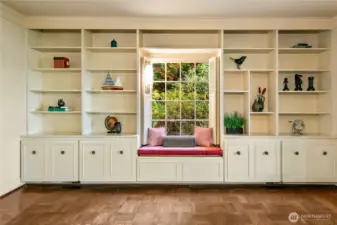 This charming built-in library wall combines timeless craftsmanship with thoughtful functionality, offering abundant storage and display space for books, art, and cherished objects. The cozy window seat, nestled beneath a multi-paned window, creates a quiet nook perfect for reading or daydreaming as soft natural light filters in through surrounding evergreens. It's a peaceful corner that captures the home’s blend of elegance and intimacy.