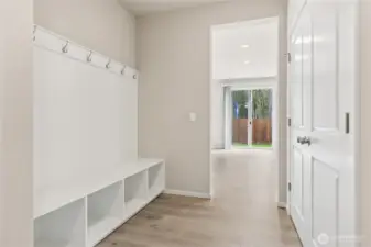 Functional mudroom drop zone with a built-in white bench with open storage cubbies below, wall-mounted coat hooks, and a view down the hallway to the sliding glass back door. Photos are representative only and may display different color package and/or upgrades.