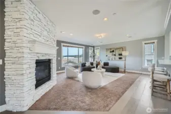 This beautifully staged living room features a modern design with a striking white stone fireplace as the focal point. Large windows fill the space with natural light and offer scenic views, while the soft gray walls and plush carpet create a warm and inviting atmosphere. Stylish furniture, including a curved sectional and two contemporary swivel chairs, is arranged around a chic round coffee table, perfectly balancing comfort and elegance.