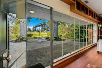 A continuous span of floor-to-ceiling commercial steel windows runs the length of the Great Room, opening directly to the Lanai and the landscaped grounds. The glazing draws in steady natural light and frames the heritage oak as a central element of the home’s indoor–outdoor rhythm.