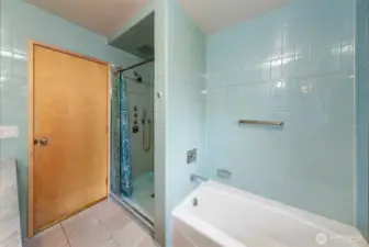 A calm, light-filled bathroom finished in soft blue tile features a walk-in shower with glass enclosure and a soaking tub set into a clean, modern surround. Warm wood accents and simple lines give the space a quiet, functional clarity.