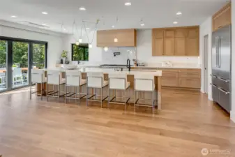A bright, modern kitchen designed for both cooking and gathering, featuring a massive slab-quartz island with a sleek waterfall edge and seating for six. Warm wood cabinetry, high-end appliances, and wide-open flow to the deck create an easy, stylish space for everyday living and effortless entertaining.