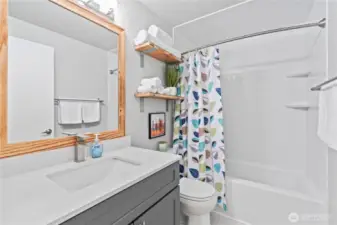 Beautifully updated full bathroom featuring a sleek white quartz vanity, modern fixtures, and a tub/shower combo with built-in shelving. Open wood shelving adds charm and functionality, while fresh paint and contemporary lighting complete the clean, inviting look.