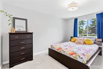 Spacious and sunlit bedroom featuring a large window with peaceful neighborhood views, plush carpeting, and a stylish modern bed with a bold floral motif. Crisp white walls and dark wood furniture offer a clean, contemporary feel—ideal as a primary bedroom or guest retreat.