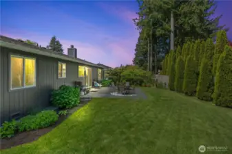 Enjoy peaceful evenings in this fully fenced backyard oasis featuring a spacious patio perfect for grilling, outdoor dining, and cozy gatherings. Surrounded by mature landscaping and tall evergreens for added privacy, this twilight view highlights the seamless indoor-outdoor lifestyle this home offers.