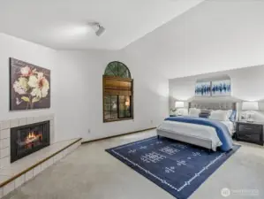 Expansive upper primary suite w vaulted ceilings, a cozy fireplace, & a signature arched window framing lush Pacific Northwest greenery