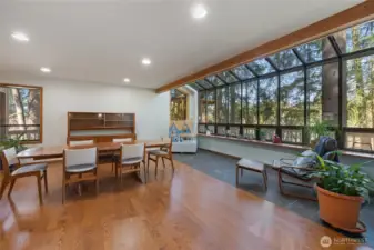 Expansive dining and sitting area with rich hardwood floors and a wall of windows capturing tranquil tree-lined views, creating a bright, inviting atmosphere.